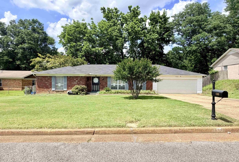 Brick-front ranch style home featuring a two-car garage, mature landscaping, a dark shingle roof, and a front entry door with a storm door