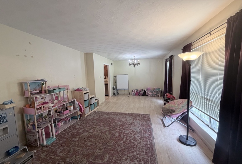 Spacious room featuring light wood-finish flooring, a decorative chandelier, and a large window with horizontal blinds and dark curtains