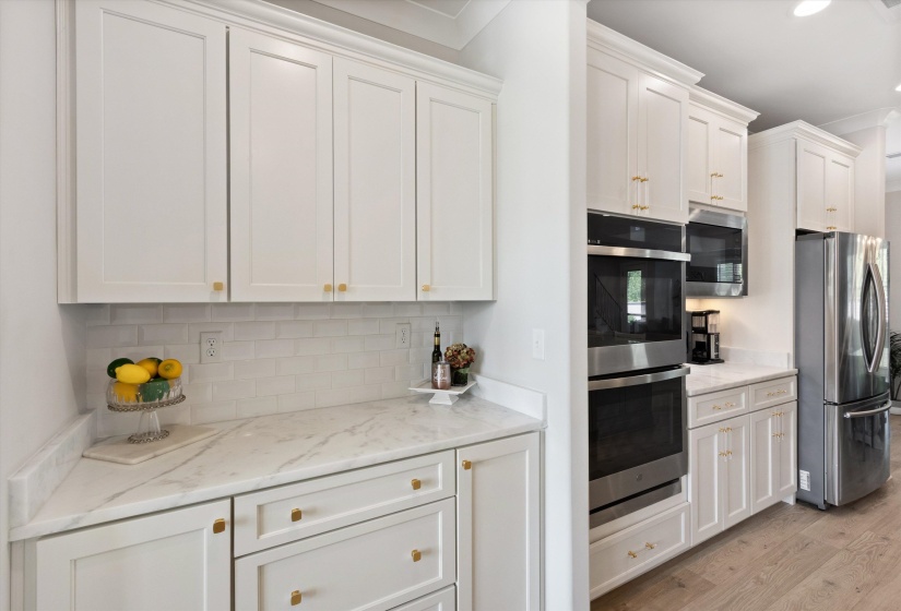 Kitchen featuring white shaker-style cabinetry with gold hardware, light-toned countertops, a white subway tile backsplash, and stainless steel double ovens