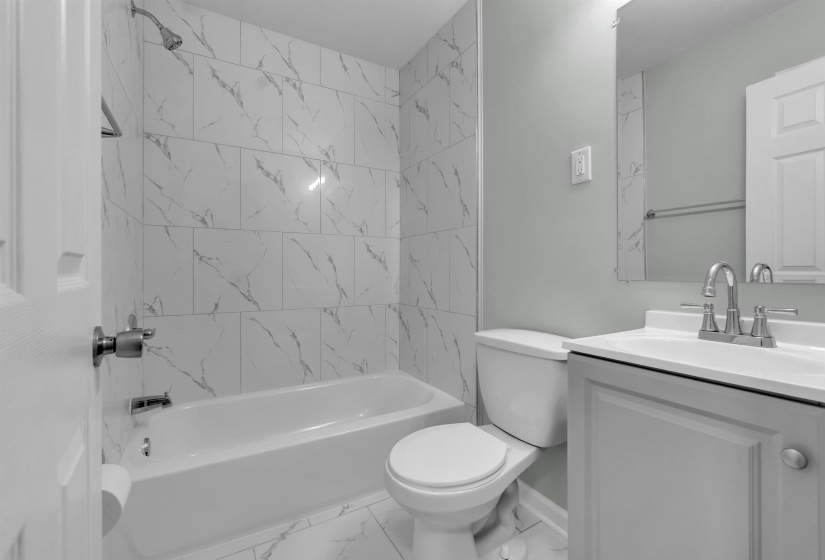 Bathroom featuring a white bathtub, marble-patterned tile shower surround, matching tile flooring, a single vanity with a white countertop, and a wall-mounted mirror