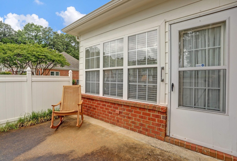 Exterior patio area featuring a white vinyl privacy fence, red brick foundation, and multi-pane windows with blinds