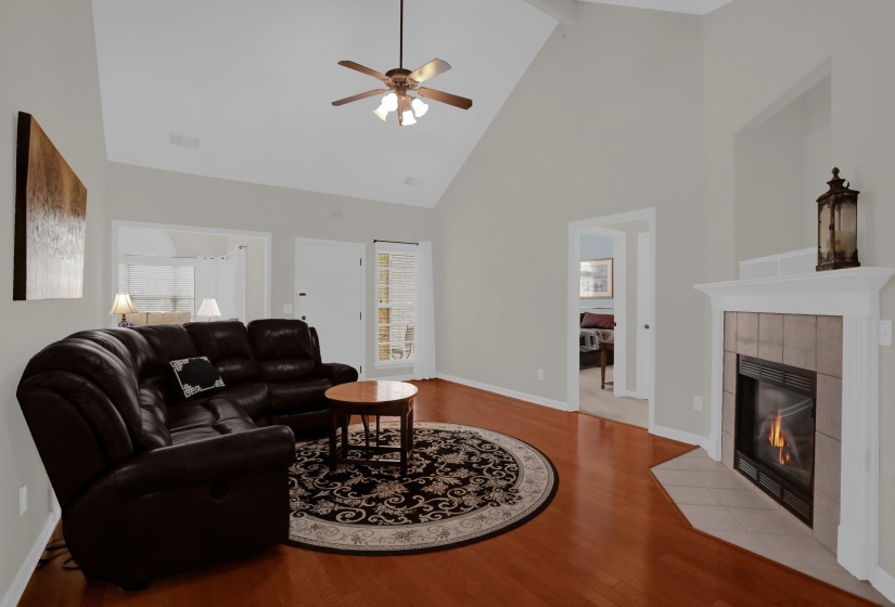 Living area featuring vaulted ceilings, wood-finish flooring, a ceiling fan with integrated lighting, a tiled fireplace with white mantel, and a doorway opening to an outdoor area