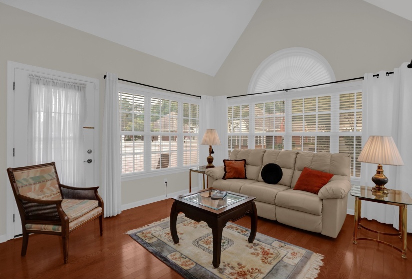 Bright living area featuring vaulted ceilings, wood-finish flooring, multiple paneled windows with blinds, and an arched transom window