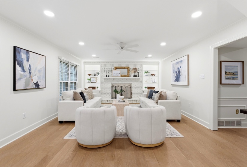 Spacious living area featuring light wood-finish flooring, a white brick fireplace with a mantel, built-in shelving, recessed lighting, and a ceiling fan