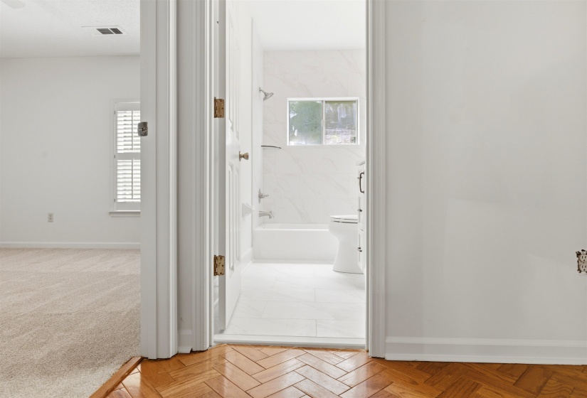Herringbone wood-finish flooring transitions to a white-tiled bathroom featuring a bathtub with a shower, a toilet, a window, and white wall tiling