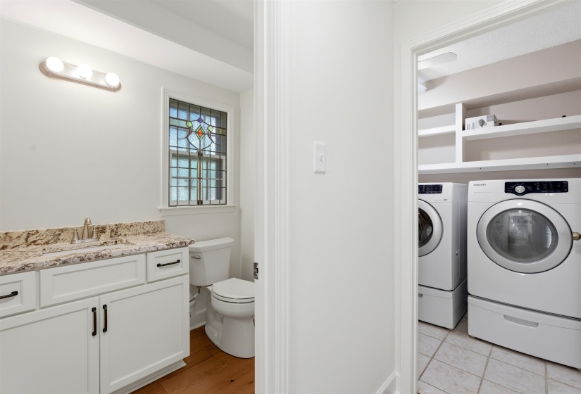 Full-sized laundry machines with built-in shelving, contrasting with a nearby vanity featuring a stone countertop, an undermount sink, and a stained glass window
