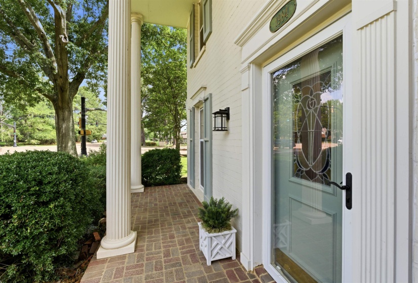 Front porch featuring brick pavers, fluted columns, a decorative glass entry door, and exterior sconce lighting
