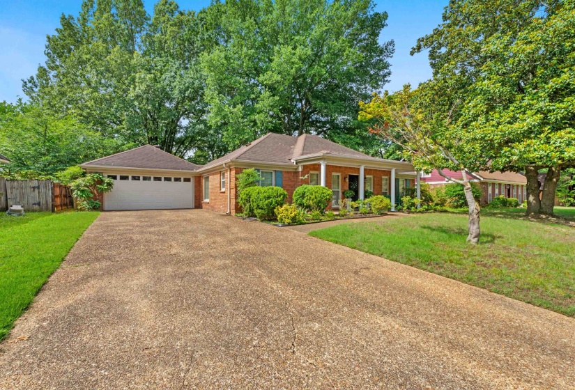 Brick exterior featuring a covered entry with white columns, a two-car garage, mature trees, and a concrete driveway