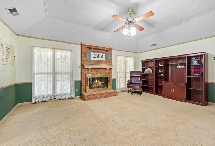 Spacious room featuring a prominent brick fireplace with a mantle, two large windows, a built-in wood-finish shelving unit, a ceiling fan with light fixture, and vaulted ceilings