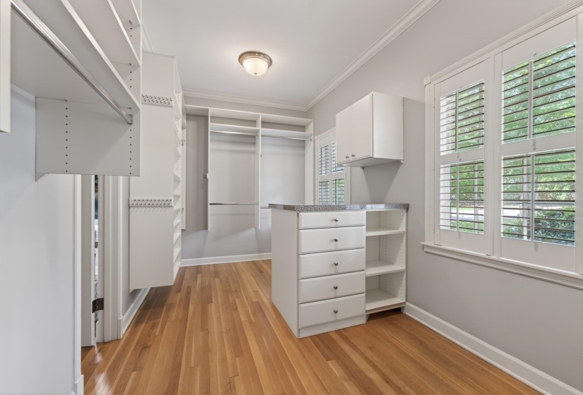 Custom closet system featuring wood-finish flooring, white plantation shutters, built-in cabinetry, and a central island with a stone-finish countertop