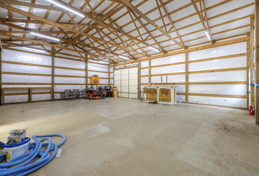 Spacious outbuilding featuring a concrete floor, exposed wood beam construction, insulated walls, fluorescent lighting, and a large roll-up door