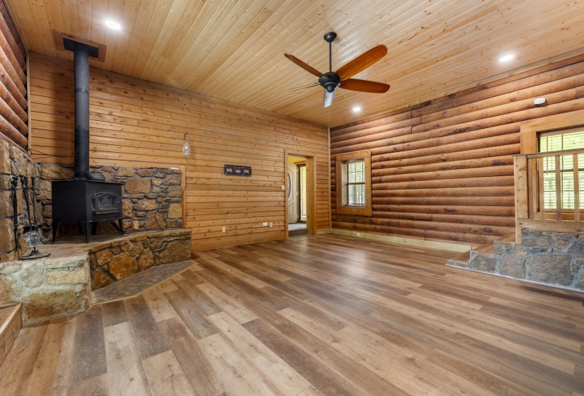 Spacious room featuring log and wood-paneled walls, wood-finish flooring, a stone-surrounded wood-burning stove, and a ceiling fan with integrated lighting