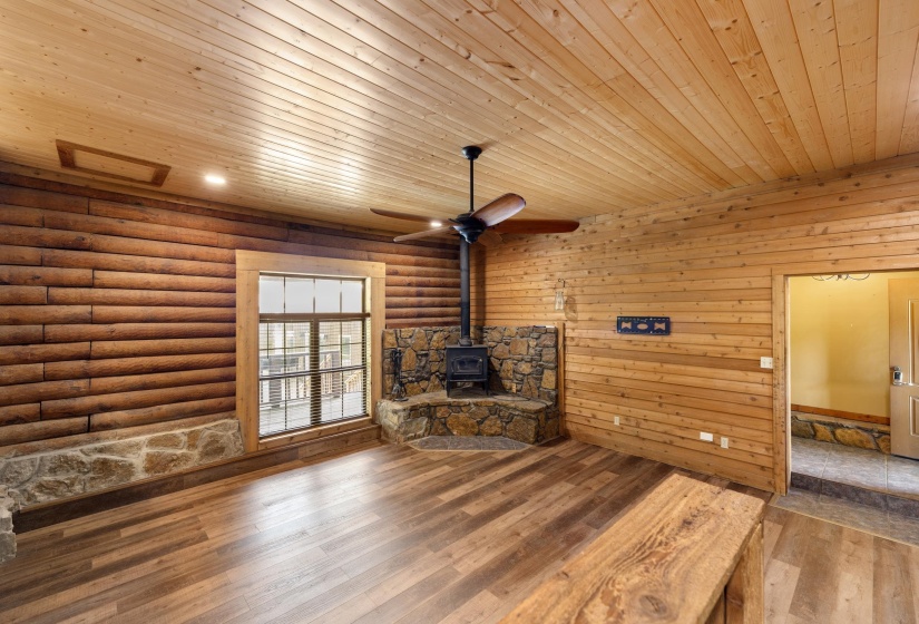 Stone-front wood-burning stove with a natural stone hearth, wood-finish flooring, wood plank ceiling, exposed log walls, and ceiling fan