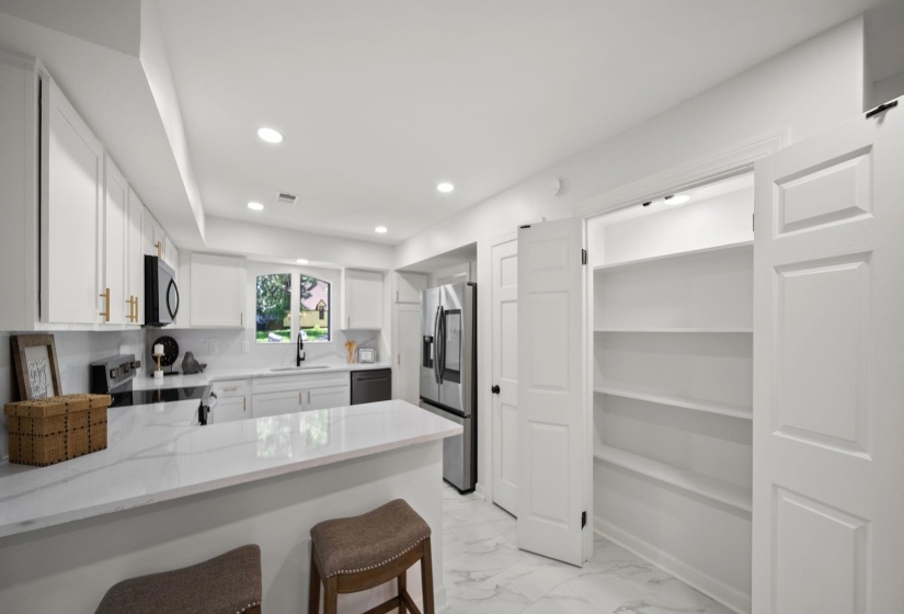 Contemporary white kitchen featuring a peninsula with a stone-finish countertop, recessed lighting, and a double-door pantry with shelving
