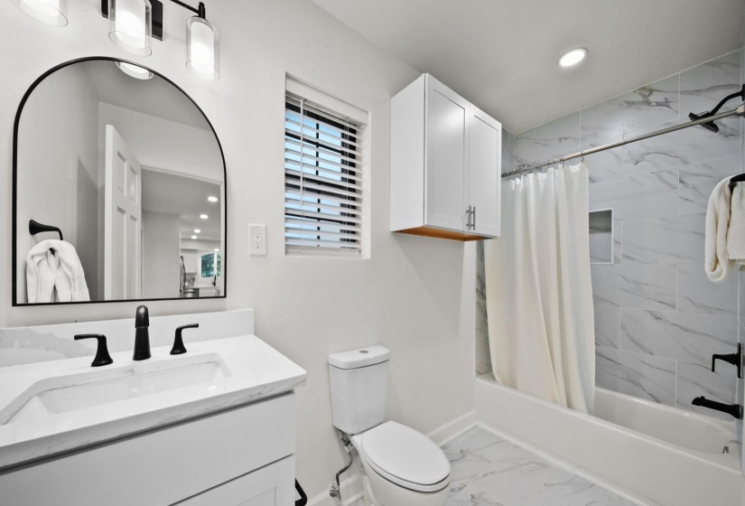 Modern bathroom featuring a vanity with a white countertop and matte black faucet, an arched mirror, a toilet, and a bathtub with white and gray tiled walls