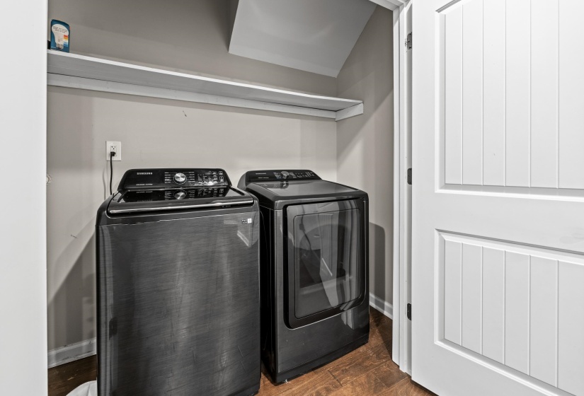 Dedicated laundry closet with wood-finish flooring, featuring a top-load washing machine, front-load dryer, and a single wall-mounted shelf