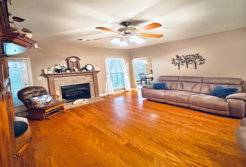 Living area featuring wood-finish flooring, a fireplace with a tiled surround and mantel, a ceiling fan with integrated lighting, and a doorway to an additional room