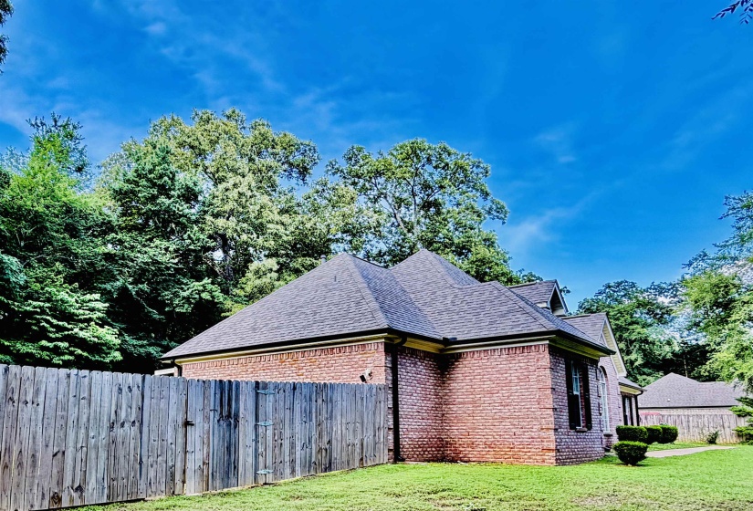 Brick exterior with black asphalt shingle roof, surrounded by mature trees, featuring a wooden privacy fence, manicured lawn, and decorative shrubbery