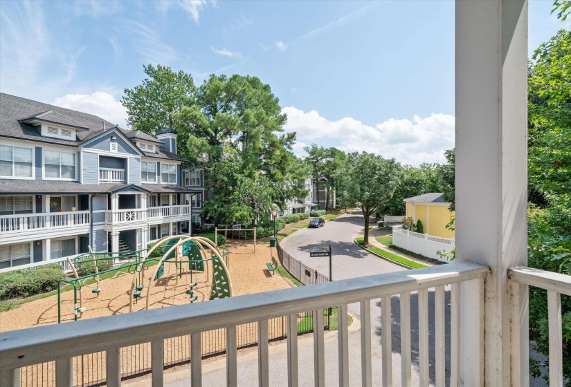 Balcony overlooking a residential community featuring multi-story buildings with blue siding and white trim, mature trees, a dedicated playground area, a paved street, and a white fence