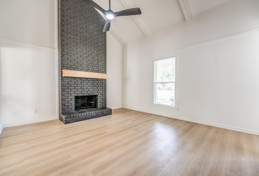Living area featuring a floor-to-ceiling dark brick fireplace with a wood mantle, vaulted ceilings with exposed beams, wood-finish flooring, and a ceiling fan
