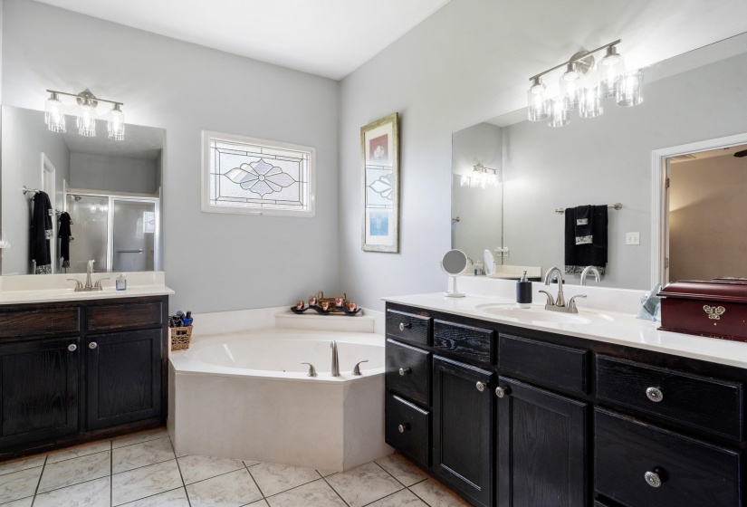 Soaking tub with a decorative privacy window, dual vanities featuring dark cabinetry, and tile flooring