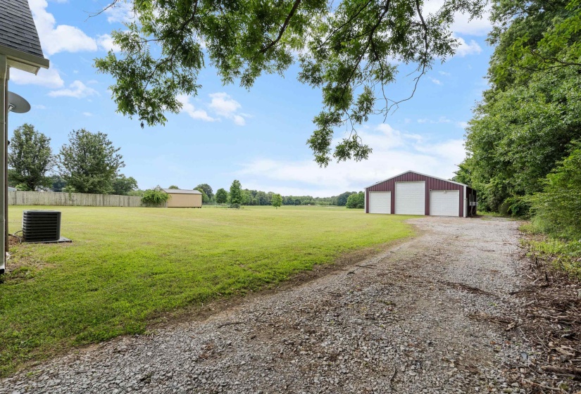 Expansive gravel driveway leading to a three-bay metal outbuilding with roll-up doors, positioned on a large grass lot