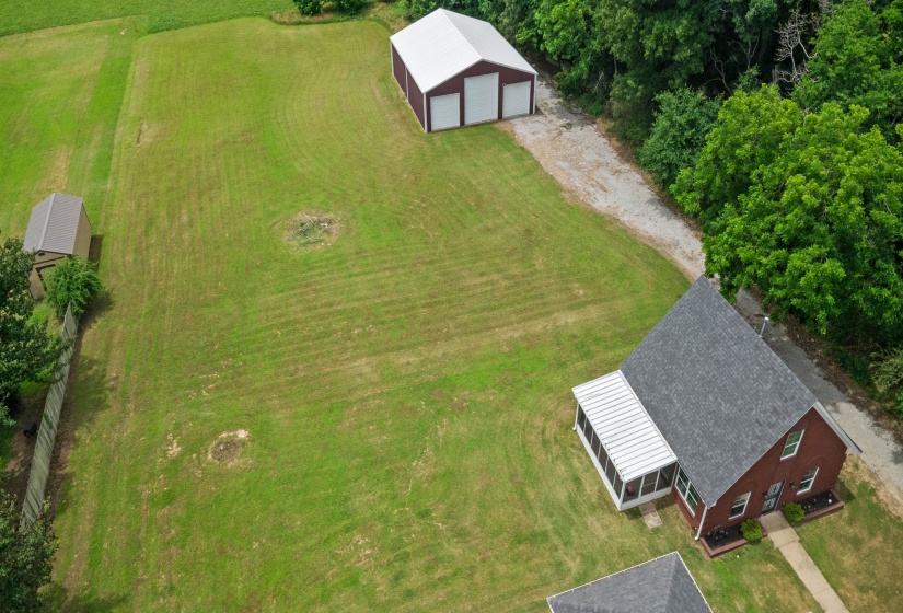 Expansive green landscape featuring a red brick residence with a gray shingle roof, an attached screened porch, a detached metal barn with three bays, and a secondary shed structure
