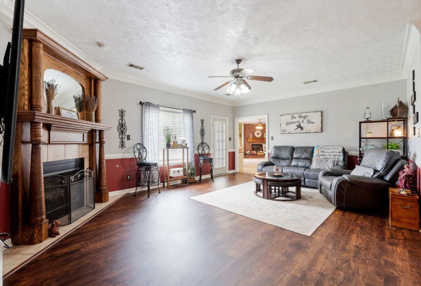 Spacious living area featuring wood-finish flooring, a traditional wood mantel fireplace with tile surround, a ceiling fan, crown molding, and a large window with blinds