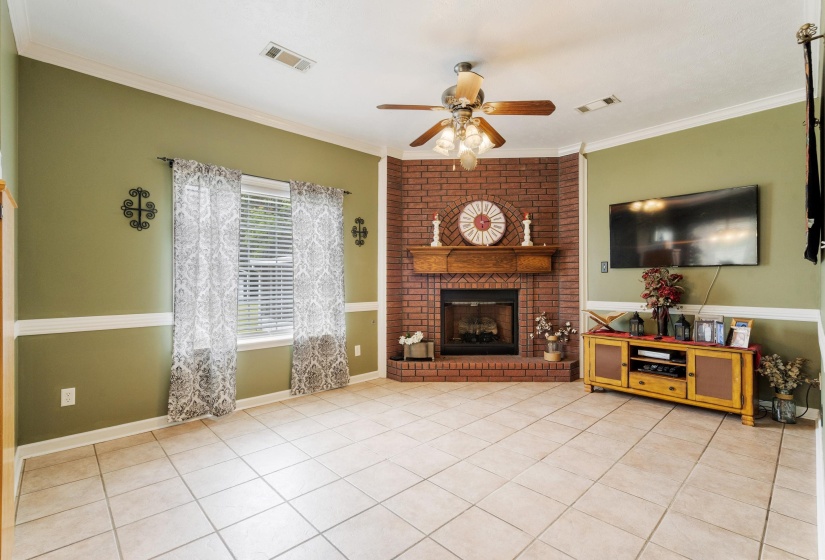 Living area featuring a corner brick fireplace with a wood mantel, tile flooring, crown molding, and a ceiling fan with integrated lighting