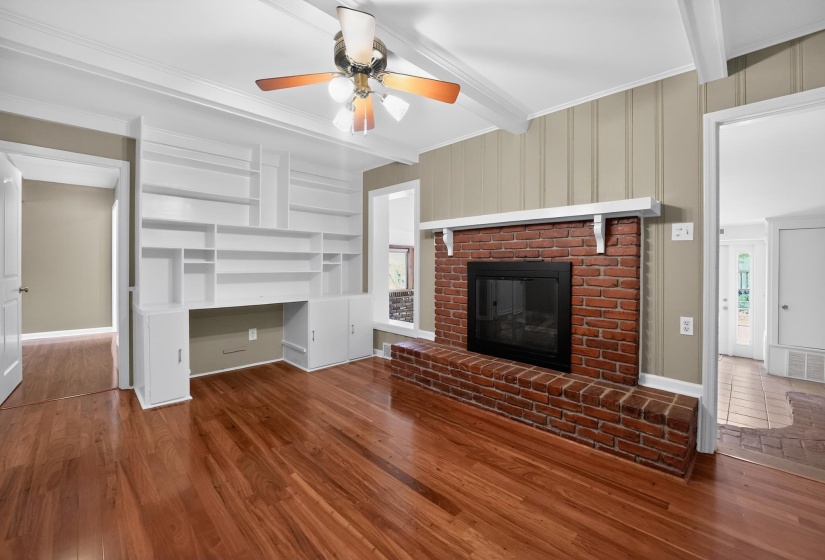 Hardwood flooring throughout featuring a brick fireplace with mantel, built-in shelving, exposed ceiling beams, and a ceiling fan