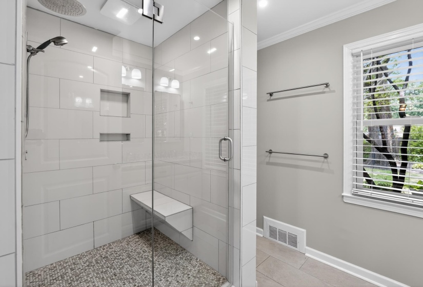 Spacious walk-in shower featuring a glass enclosure, pebble-tile floor, built-in bench, recessed wall niches, and overhead rainfall showerhead