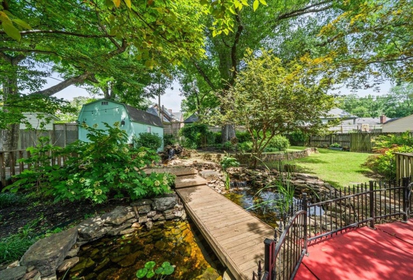 Lush backyard oasis featuring a pond with a wooden bridge, a red-painted deck, a light blue storage shed, mature trees, and natural stone landscaping