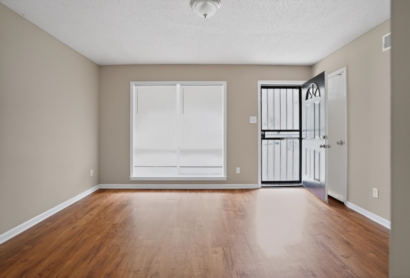 Spacious room featuring wood-finish flooring, neutral-toned walls, a large window with white blinds, a ceiling-mounted light fixture, and a front entry door with an arch-top design and security screen