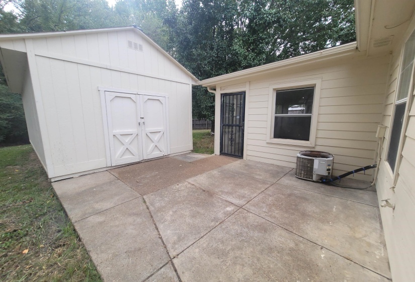 Concrete patio area featuring a detached storage shed with double doors, siding exterior, and an exterior entry door with a security grate
