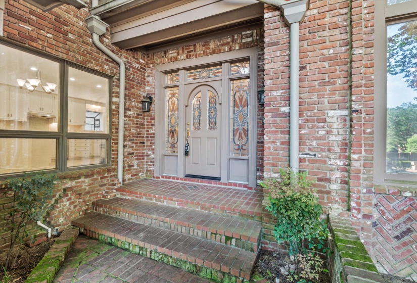 Brick exterior featuring a paneled entry door with decorative glass sidelights, a porch light fixture, and a multi-pane window revealing interior cabinetry