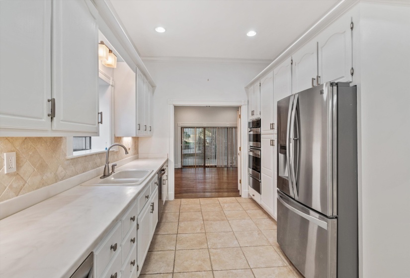 Galley kitchen featuring white cabinetry, light-toned countertops, a tile backsplash, and stainless steel appliances including a refrigerator and double ovens