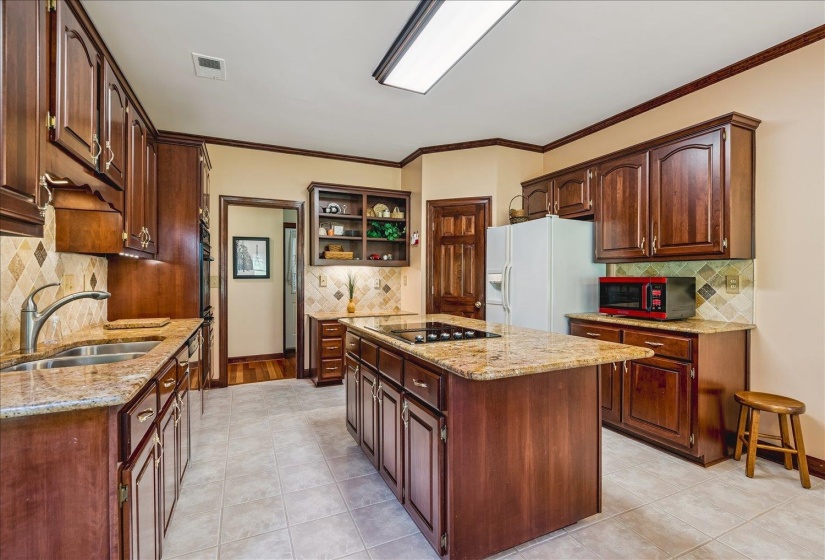 Kitchen featuring rich wood cabinetry, granite countertops, tile backsplash, a center island with a cooktop, and neutral tile flooring