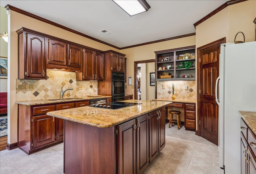 Kitchen featuring a center island with integrated cooktop, granite countertops, wood cabinetry, tile backsplash, and built-in desk area