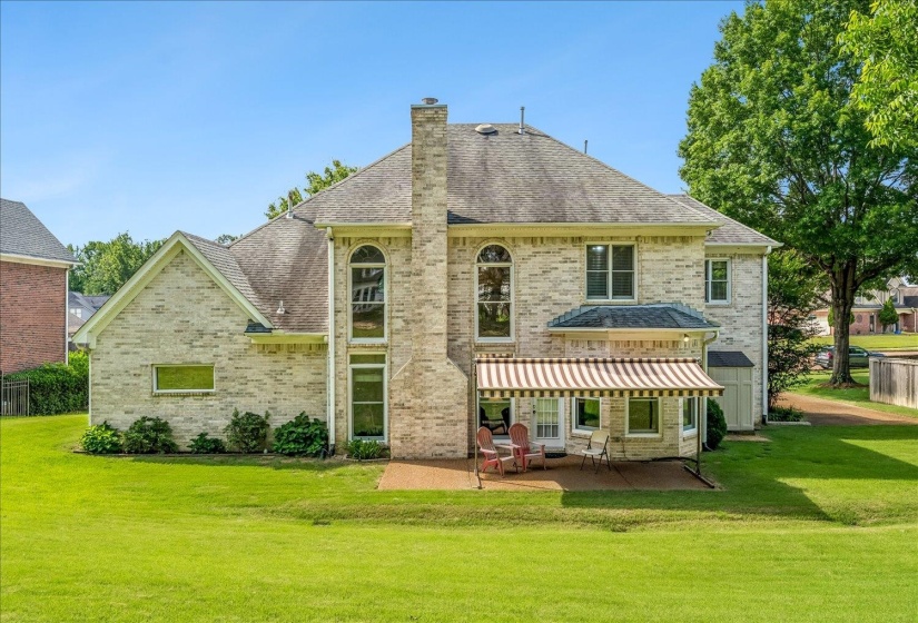 Spacious backyard featuring a large brick exterior, a patio with a striped awning, a prominent brick chimney, and mature trees