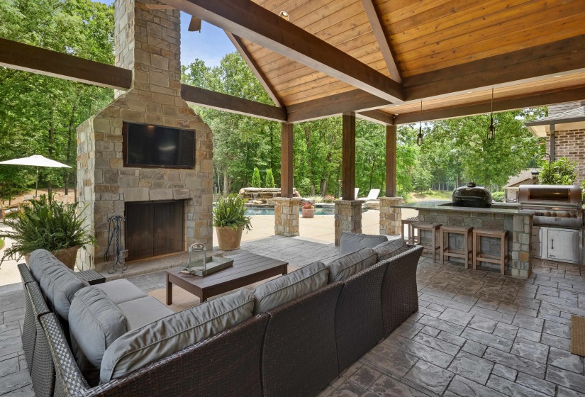 Expansive covered patio featuring a stone fireplace with integrated television, an outdoor kitchen with built-in grill and refrigerator, wood-plank cathedral ceiling, and stamped concrete flooring
