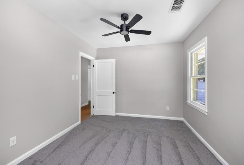 Neutral gray wall finish with a white ceiling, featuring gray carpeting, a multi-pane window with white trim, a white paneled door with black hardware, and a black ceiling fan