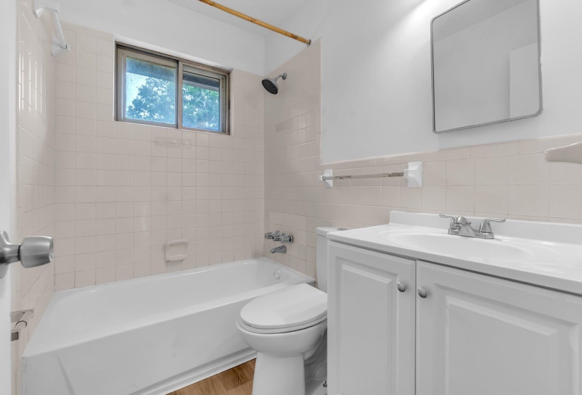 Bathroom featuring a white vanity with built-in sink, a rectangular framed mirror, an integrated tub and shower, a window, and wood-finish flooring