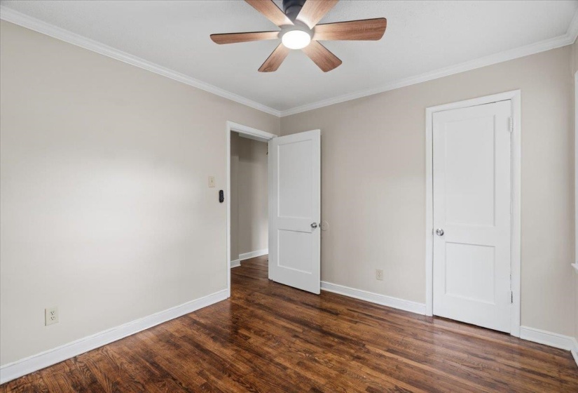 Room featuring rich wood-finish flooring, light neutral-toned walls, white crown molding, and a ceiling fan with light fixture