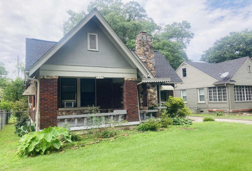 Gabled bungalow featuring a covered porch with brick and stone accents, a prominent stone chimney, and a dark shingle roof