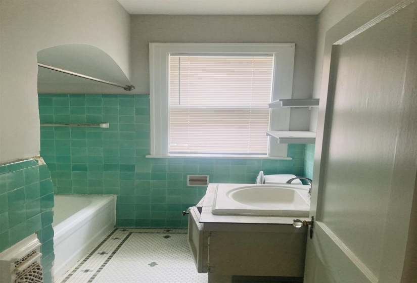 Bathroom featuring a window with blinds, a vanity with an integrated sink, built-in shelving, green square wall tiles, and white hexagonal floor tiles with dark accents