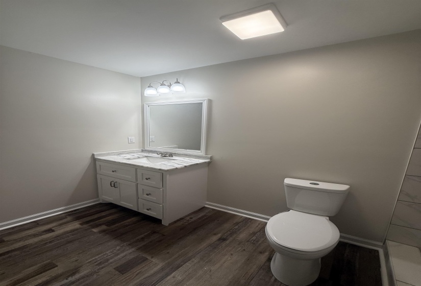 Bathroom featuring a white vanity with a marble-patterned countertop, wood-finish flooring, and a contemporary wall-mounted light fixture