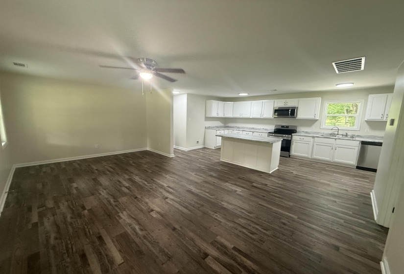 Open-concept kitchen and living area featuring wood-finish flooring, white cabinetry, a kitchen island with stone-look countertop, stainless steel appliances, and a ceiling fan