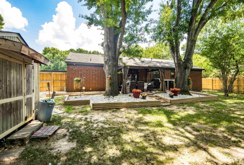Expansive backyard featuring a brick exterior, privacy fencing, a covered patio area, raised garden beds with white stone accents, and mature trees