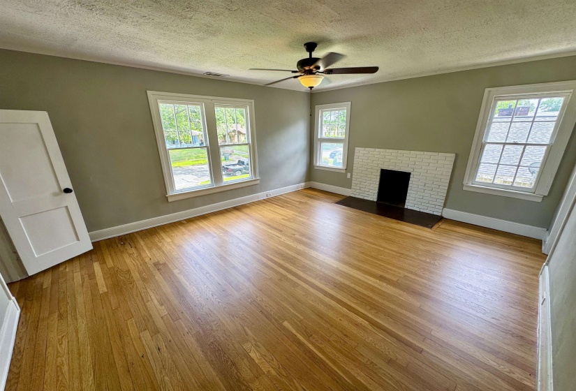 Room featuring natural wood flooring, a white-painted brick fireplace, multiple windows with white trim, and a ceiling fan with integrated lighting