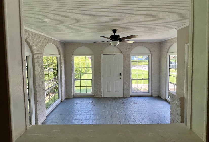 Enclosed porch featuring a brick-patterned floor, white painted brick walls, arched window openings with gridded windows, a white entry door, and a ceiling fan